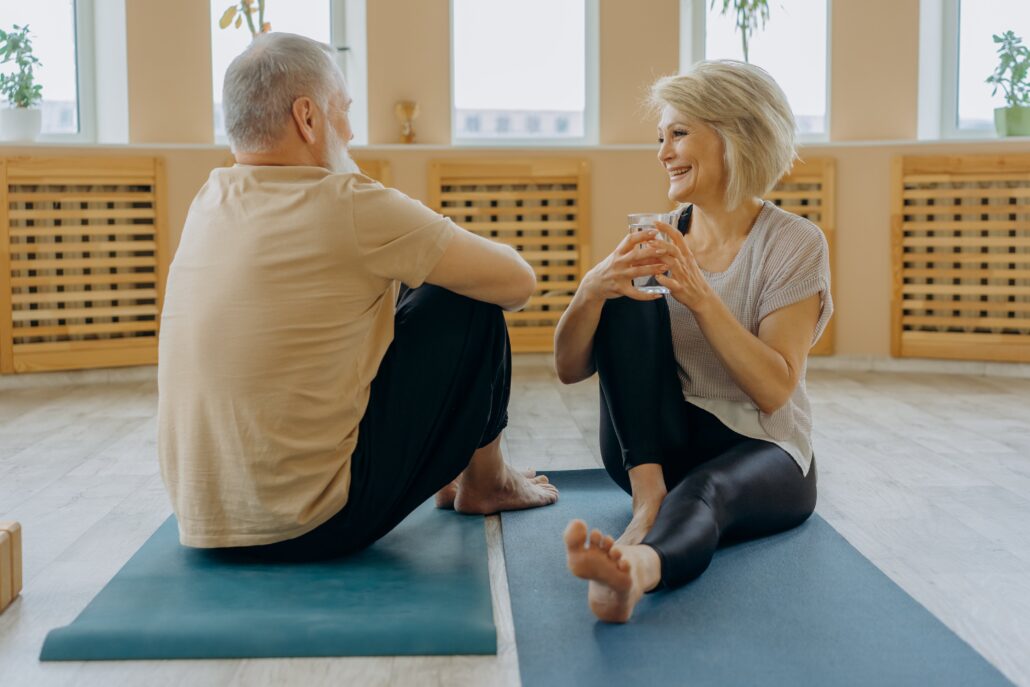 Two older yoga practitioners looking at each other sitting on their mats in a yoga studio.